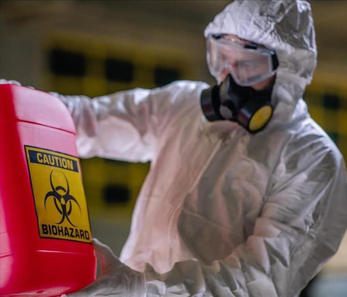 A man dressed in a white hazmat suit and holding a red bio-hazard container.